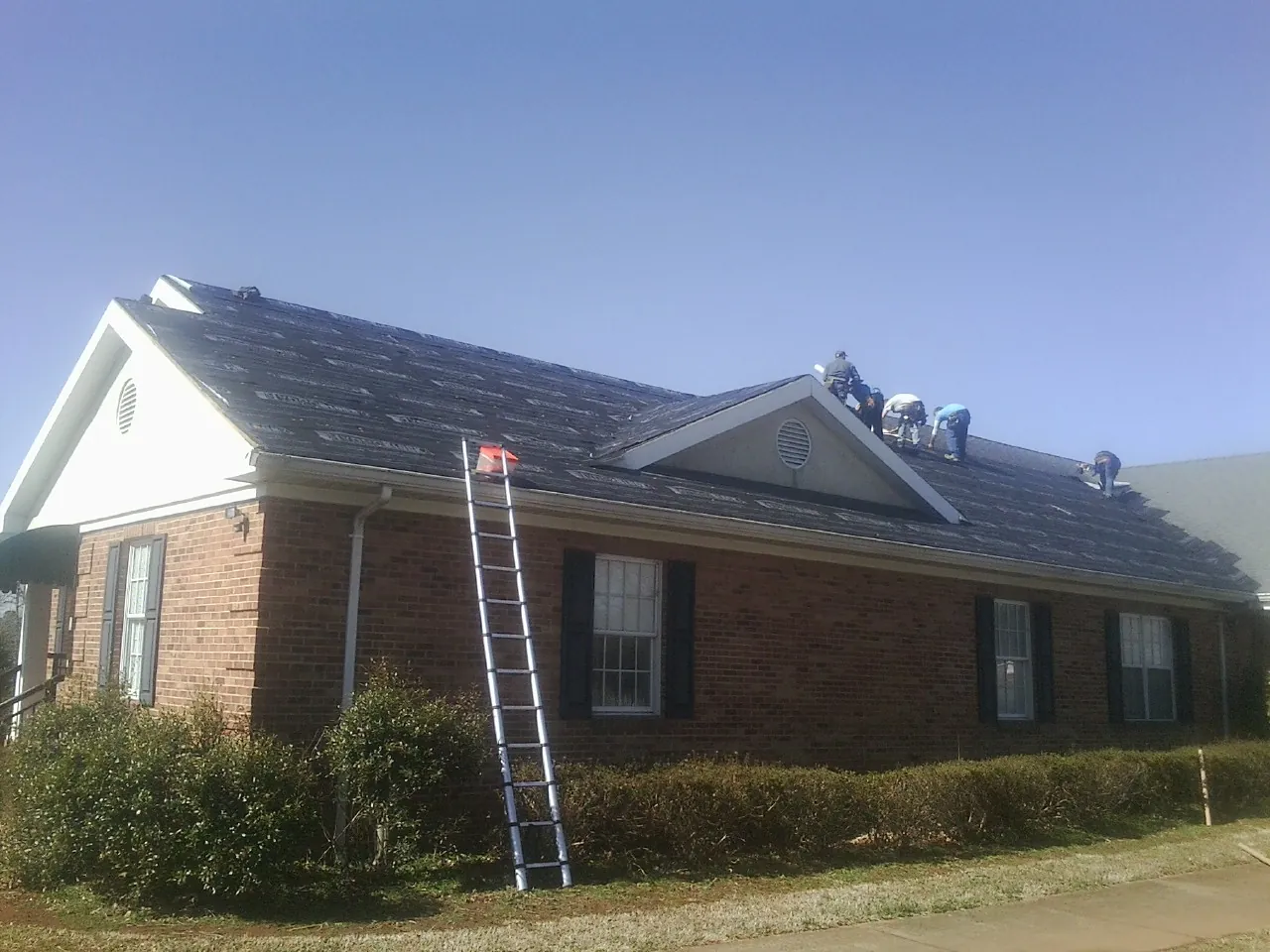 Crew tearing shingles off a brick ranch roof with a ladder set — Metal Roof Repair work by our crew in Ripley