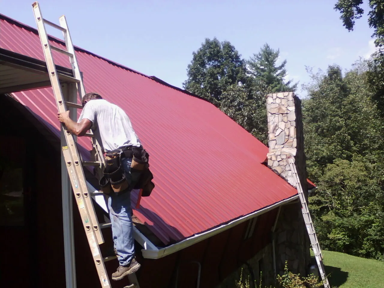 Roofer on a ladder beside a red metal roof and stone chimney — Soffit & Fascia Repair by Roofing Brothers in Ripley